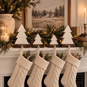 May include: Four cream-colored cable knit stockings hang from a mantelpiece, each suspended by a wooden Christmas tree-shaped stocking holder. The stockings feature a textured knit pattern and are filled with small sprigs of greenery.