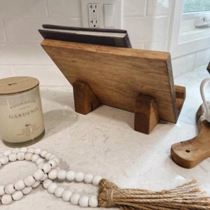 May include: A wooden cookbook stand with a book on top. The stand has a natural wood finish and is made of solid wood. The stand is sitting on a white countertop.