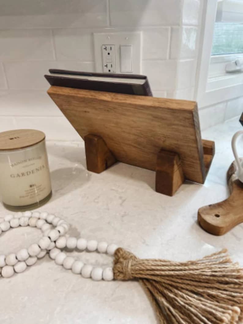 May include: A wooden cookbook stand with a book on top. The stand has a natural wood finish and is made of solid wood. The stand is sitting on a white countertop.