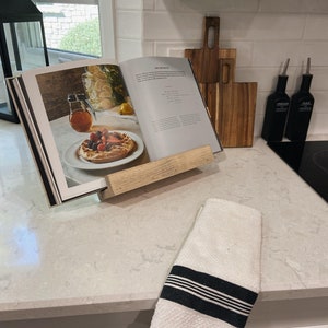 May include: A white wooden cookbook stand with a cookbook open on top. The cookbook has a picture of a dessert with fruit and whipped cream. There are two wooden cutting boards and two glass bottles with labels on the counter behind the stand.