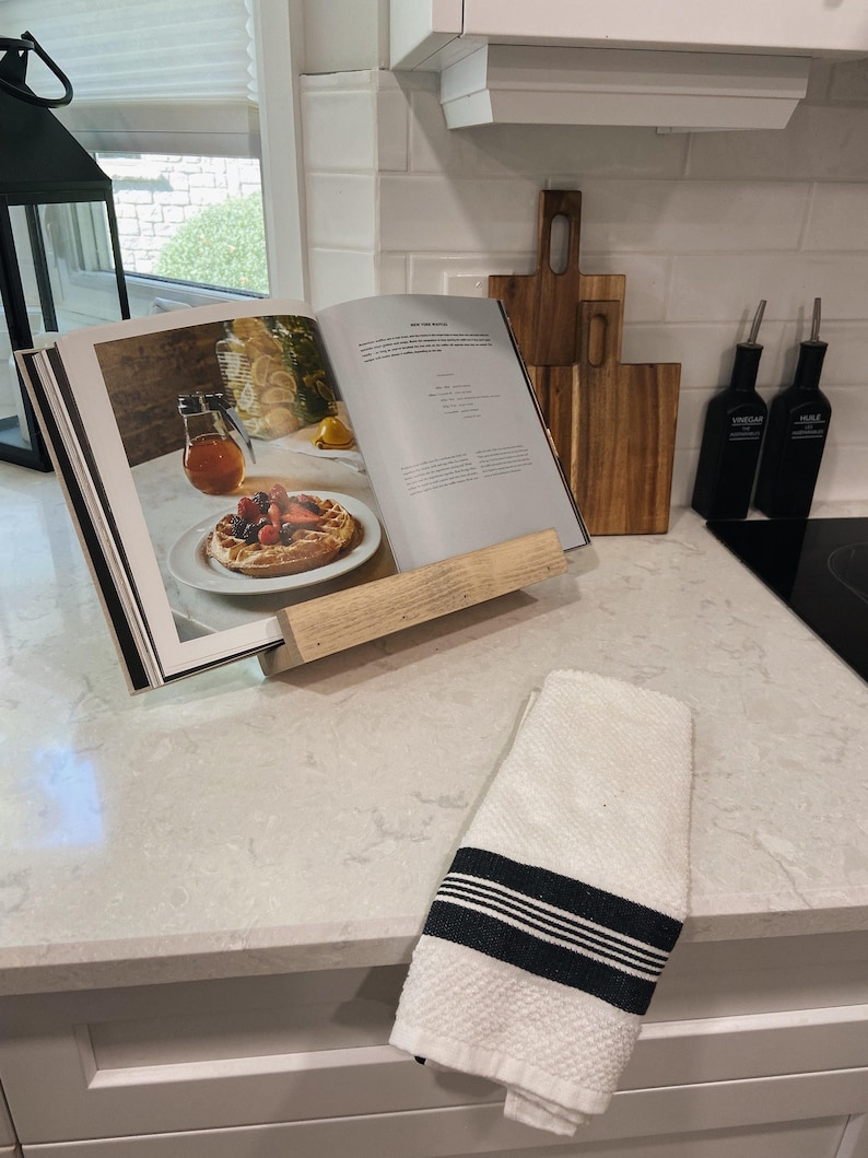 May include: A white wooden cookbook stand with a cookbook open on top. The cookbook has a picture of a dessert with fruit and whipped cream. There are two wooden cutting boards and two glass bottles with labels on the counter behind the stand.
