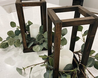 May include: Two wooden candle lanterns with white candles inside, decorated with green eucalyptus greenery.