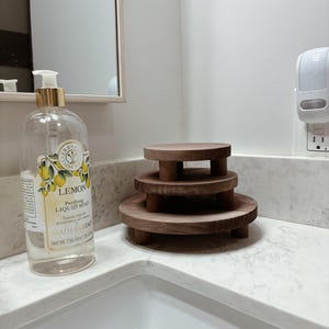 以下が含まれることがあります： バスルームに置かれた3段のウッドトレイ。トレイはダークウッド製で、ナチュラルな仕上げ。一番上のトレイが最も小さく、一番下のトレイが最も大きい。トレイは積み重ねられており、白いカウンタートップの上に置かれている。