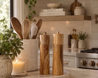 May include: Two cylindrical wooden salt and pepper grinders with metallic top knobs, positioned on a circular stone tray alongside a ceramic utensil crock containing wooden spoons, a whisk, and a spatula, plus a lit candle in a glass jar.