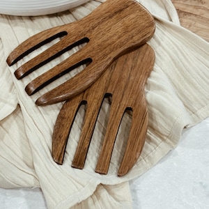 Puede incluir: Dos servidores de ensalada de madera con un acabado marrón. Los servidores tienen cuatro púas cada uno y están hechos de madera natural.