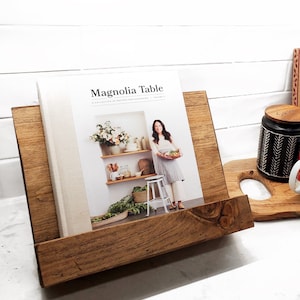 May include: A wooden cookbook stand with a brown finish holds a cookbook titled "Magnolia Table: A Collection of Recipes for Gathering, Volume 1". The cookbook features a photo of a woman holding a bowl of food.