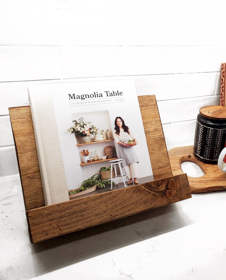 May include: A wooden cookbook stand with a brown finish holds a cookbook titled "Magnolia Table: A Collection of Recipes for Gathering, Volume 1". The cookbook features a photo of a woman holding a bowl of food.