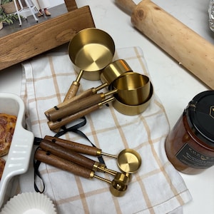 May include: A set of gold measuring cups and spoons with wooden handles. The cups and spoons are arranged on a white and brown checkered tablecloth. The cups are nested inside each other, and the spoons are laid out next to them. The set is perfect for baking or cooking.