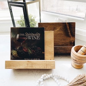 May include: A wooden book stand with a book titled "The Spirituality of Wine" by Tom Harpur. The book stand is made of natural wood and has a rustic look. There is a white beaded garland with a brown tassel in the foreground.