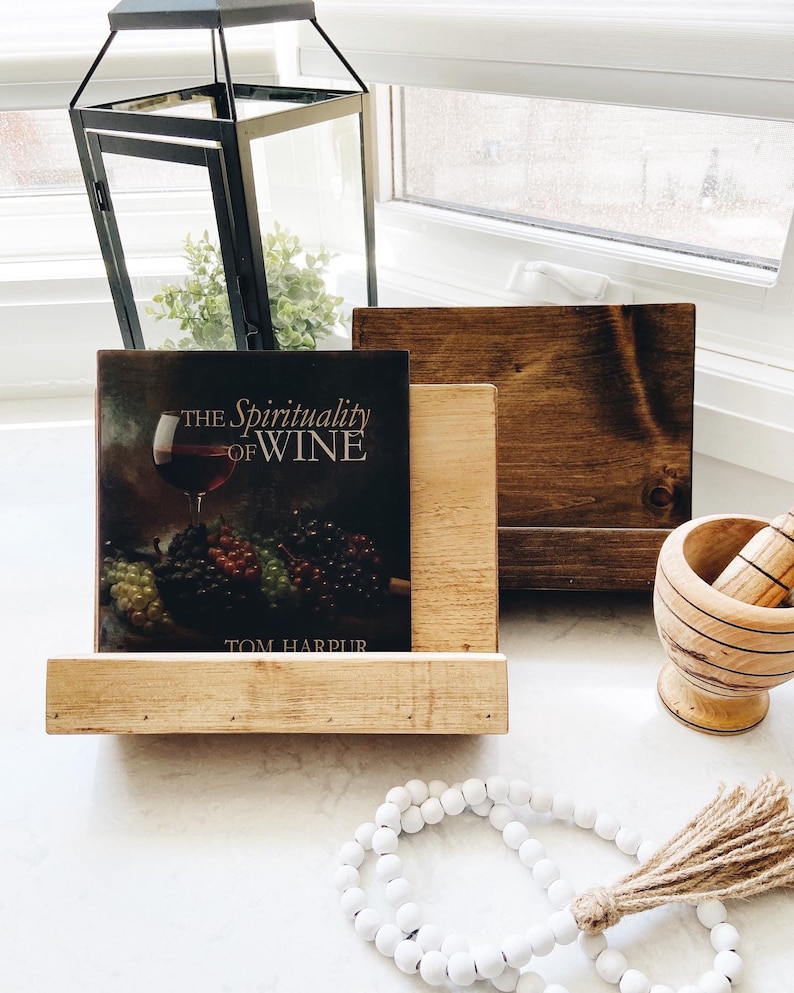 May include: A wooden book stand with a book titled "The Spirituality of Wine" by Tom Harpur. The book stand is made of natural wood and has a rustic look. There is a white beaded garland with a brown tassel in the foreground.