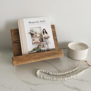 May include: A wooden book stand with a cookbook titled "Magnolia Table" on top. The cookbook features a woman holding a bowl of produce. A white candle and a string of white beads are on the surface below the stand.