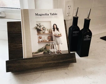 May include: A dark brown wooden cookbook stand holding a cookbook titled "Magnolia Table" with a white background and a woman holding a plate of food. Two black glass bottles with silver tops are on the counter, one labeled "Vinegar" and the other "Oil".