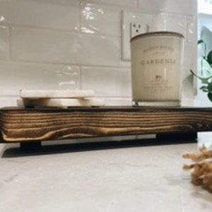 May include: A rustic wooden tray with a dark brown stain and black metal legs. The tray is holding a white candle with the word "GARDENIA" printed on the label and two white marble coasters.