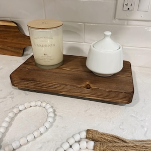 Puede incluir: Una bandeja de madera con una vela blanca y un azucarero blanco. La vela está etiquetada como "Maison Bougie Collection Gardenia". La bandeja está sobre una encimera blanca con una guirnalda de cuentas blancas con borlas marrones.