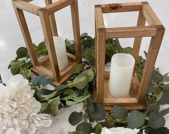 May include: Two wooden candle lanterns with white candles inside. The lanterns are surrounded by greenery and white flowers.