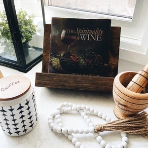 May include: A white ceramic jar with a black and white geometric pattern, labeled "Coffee", sits on a white countertop next to a wooden book stand holding a book titled "The Spirituality of Wine" by Tom Harpur. A white beaded garland with a brown tassel and a wooden mortar and pestle are also on the countertop.