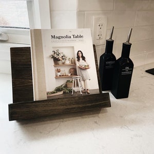 May include: A cookbook titled "Magnolia Table" by Joanna Gaines, volume 2, with a white cover and a photo of a woman holding a plate of food. The cookbook is displayed in a wooden stand with two black bottles labeled "Vinegar" and "Oil" on a white countertop.