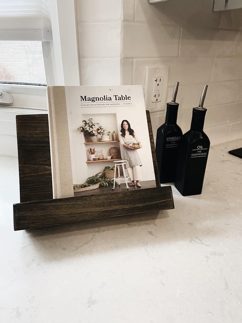 May include: A cookbook titled "Magnolia Table" by Joanna Gaines, volume 2, with a white cover and a photo of a woman holding a plate of food. The cookbook is displayed in a wooden stand with two black bottles labeled "Vinegar" and "Oil" on a white countertop.