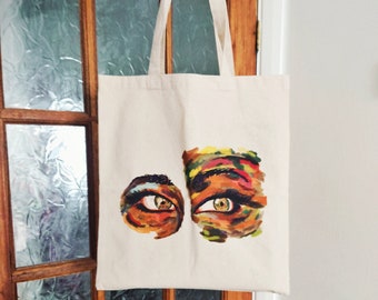 May include: A white canvas tote bag featuring a colourful painting of two eyes on the front. The eyes are painted in shades of orange, yellow, red and green.