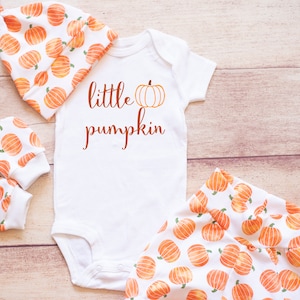 May include: A white baby outfit set with a pumpkin print. The set includes a onesie, hat, and mittens. The onesie has the text "little pumpkin" with a small pumpkin graphic.