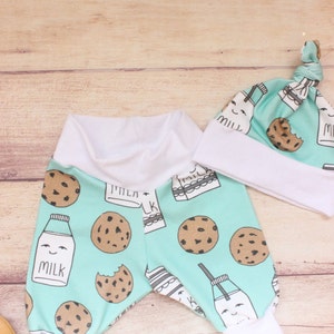 May include: A baby outfit set with a pair of blue and white milk carton and cookie print pants and a matching blue and white milk carton and cookie print hat. The hat has a white brim and a white knot on top.