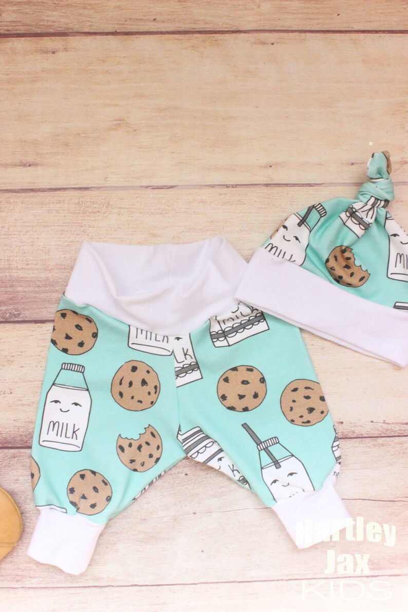 May include: A baby outfit set with a pair of blue and white milk carton and cookie print pants and a matching blue and white milk carton and cookie print hat. The hat has a white brim and a white knot on top.