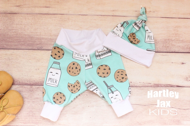 May include: A baby outfit set with a light blue and white milk carton and cookie print. The set includes a pair of pants and a hat. The hat has a knot on top. The pants have a white elastic waistband and white cuffs. The text "Hartley Jax Kids" is printed on the bottom right corner of the image.