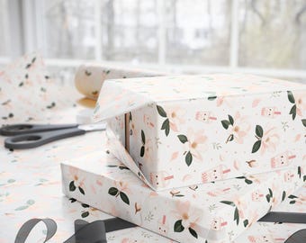May include: Two rectangular boxes wrapped in white paper featuring a repeating pattern of light pink magnolia flowers, green leaves, and small cartoon bubble tea cups with smiling faces. A black ribbon and black scissors lie on the surface.