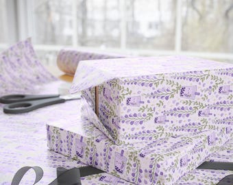 May include: Wrapping paper featuring a repeating pattern of purple wisteria flowers, green leaves, and small boba tea cups with smiling faces. The paper is used to wrap two rectangular boxes, with a roll of paper and black scissors nearby.