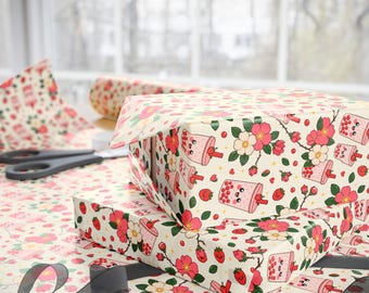 May include: Wrapping paper featuring a repeating pattern of pink boba tea cups with smiling faces, red strawberries, and pink and white flowers on a cream background. The paper is wrapped around rectangular boxes with black scissors nearby.