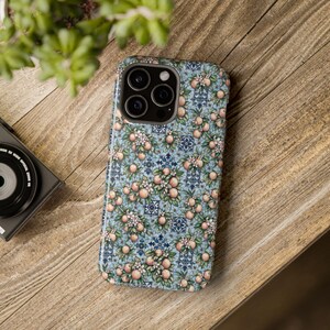 May include: A smartphone case featuring a repeating pattern of light orange citrus fruits, green leaves, and white blossoms set against a light blue background with dark blue geometric tile accents. The case has a cutout for a triple camera system.