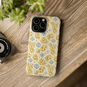 May include: A smartphone case featuring a repeating pattern of yellow lemons on green leafy branches interspersed with small blue and white decorative tile motifs against a cream-colored background, with a black camera lens array at the top.