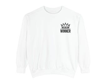 May include: A white long-sleeve sweatshirt featuring a black graphic on the upper left chest. The graphic consists of a stylized crown icon positioned above the word WINNER in bold, capital, sans-serif letters with a horizontal line.