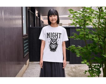 Op de afbeelding: Wit T-shirt met het woord "NIGHT" boven een gedetailleerde zwart-wit illustratie van een uil. Het uilontwerp heeft ingewikkelde veerd details en doordringende ogen. Het T-shirt is een klassieke ronde hals.