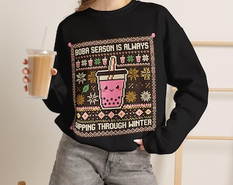 May include: A black long-sleeve sweatshirt featuring a graphic print with the text BOBA SEASON IS ALWAYS SIPPING THROUGH WINTER. The design includes a pink boba tea cup with a face, surrounded by snowflake and boba cup patterns.
