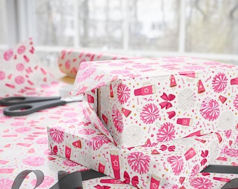 May include: Wrapping paper featuring a repeating pattern of pink cheerleading pom-poms, megaphones, bows, and tumblers on a white background. The paper is used to wrap two rectangular boxes, with a black ribbon and black scissors nearby.