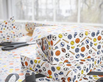 May include: Two rectangular boxes wrapped in white paper featuring a repeating pattern of colorful sports equipment including basketballs, baseballs, footballs, bats, pom-poms, and sneakers. A roll of matching wrapping paper and scissors are nearby.