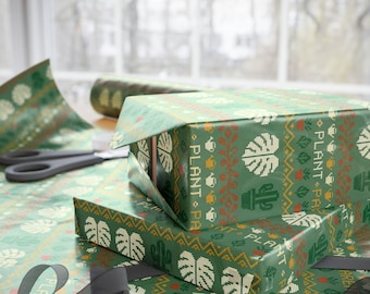 May include: Green wrapping paper featuring a repeating pattern of white monstera leaves, cacti, and watering cans with the text PLANT. The paper is wrapped around rectangular boxes, with a roll and black ribbon visible on the surface.