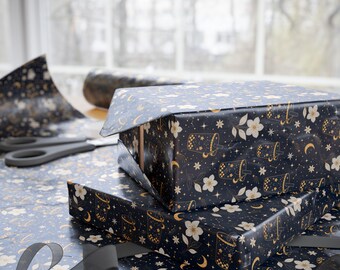 May include: A rectangular gift box wrapped in dark navy blue paper featuring a repeating pattern of gold boba tea cups, white flowers, crescent moons, and small stars. The box sits on a matching sheet of wrapping paper with a black ribbon nearby.