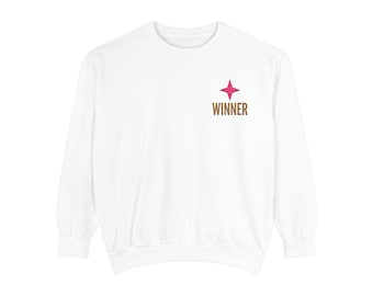 May include: A white long-sleeve sweatshirt featuring a small graphic on the upper left chest. The graphic consists of a four-pointed pink star positioned directly above the word WINNER printed in brown capital letters.