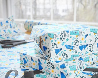 May include: White wrapping paper featuring a repeating pattern of blue swim fins, black swimming goggles, teal water bottles with the text SWIM FAST, yellow rubber ducks, orange suns, and teal starfish, with a roll and scissors nearby.