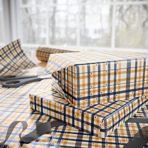 May include: Two gift boxes wrapped in plaid patterned paper with navy blue, tan, and white stripes and small red stars. The boxes are stacked on top of each other. A roll of wrapping paper and scissors are in the background.