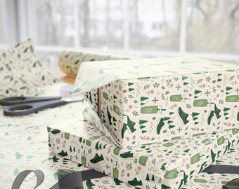 May include: Wrapping paper featuring a repeating pattern of green sneakers, tumblers with straws, pine trees, mountains, and small pink flowers on a cream background. The paper is shown wrapped around rectangular boxes with black scissors nearby.