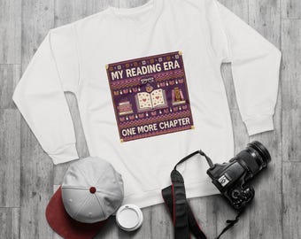 May include: A white long-sleeve sweatshirt featuring a central graphic with a purple background, a pattern of books, glasses, and a mug, with the text "MY READING ERA" and "ONE MORE CHAPTER" printed in white, accompanied by a baseball cap and a camera.
