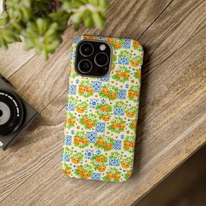 May include: A smartphone case featuring a repeating pattern of smiling orange fruits with green leaves and white blossoms, interspersed with small blue and white geometric tile motifs and tiny bees on a light cream background.