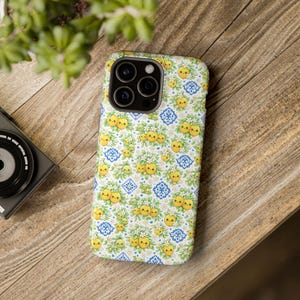 May include: A smartphone case featuring a repeating pattern of yellow cartoon lemons with green leaves, white blossoms, and blue geometric tile motifs on a white background. The case has a cutout for a triple-lens camera system on the back.