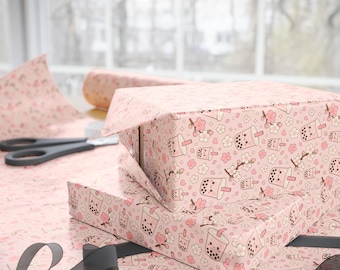 May include: Light pink wrapping paper featuring a repeating pattern of smiling bubble tea cups, cherry blossom flowers, and small hearts. The paper is shown partially wrapped around a rectangular box, with black scissors and a black ribbon nearby.