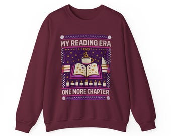 May include: A maroon crewneck sweatshirt featuring a graphic print with the text MY READING ERA and ONE MORE CHAPTER. The design includes an open book, a steaming mug, a cat, stacks of books, and candles against a starry purple background.