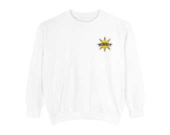 May include: A white long-sleeve sweatshirt featuring a small graphic on the upper left chest. The graphic is a yellow starburst shape with a distressed texture, containing the word WINNER printed in bold, dark blue capital letters.