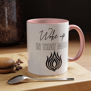 May include: White ceramic mug with a pink interior and handle. The mug features the text "Woke up IN THAT MODE" in black script and a black flame design. A silver spoon and a macaron are on a wooden surface.